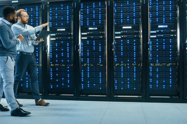 IT Technician with a Laptop Computer and Black Male Engineer Colleague are Talking in Data Center while Walking Next to Server Racks. Running Diagnostics or Doing Maintenance Work. (IT Technician with a Laptop Computer and Black Male Engineer Colleagu
